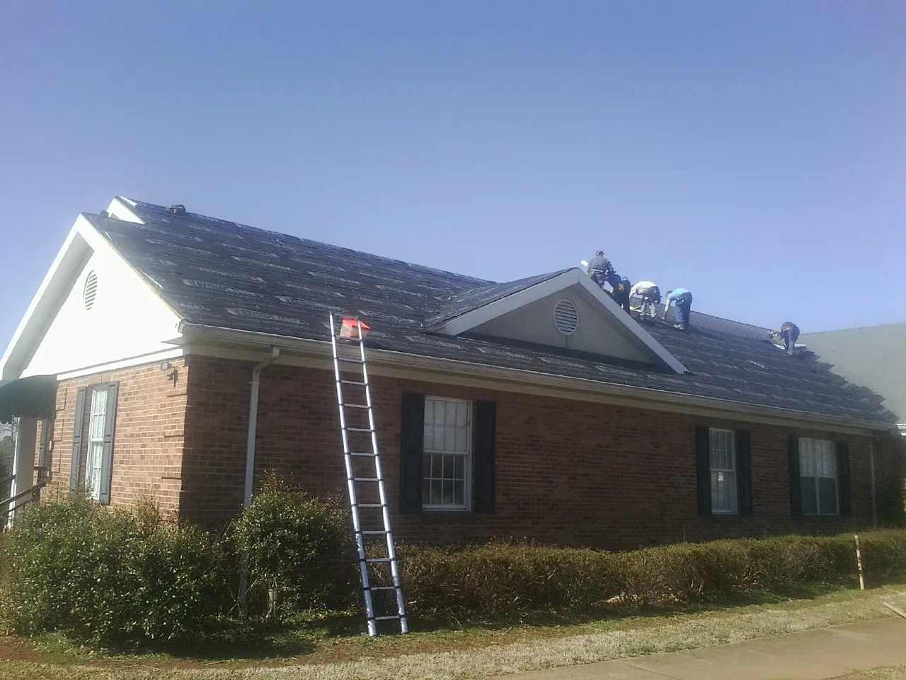 Crew tearing shingles off a brick ranch roof with a ladder set — Metal Roof Repair work by our crew in Houlka