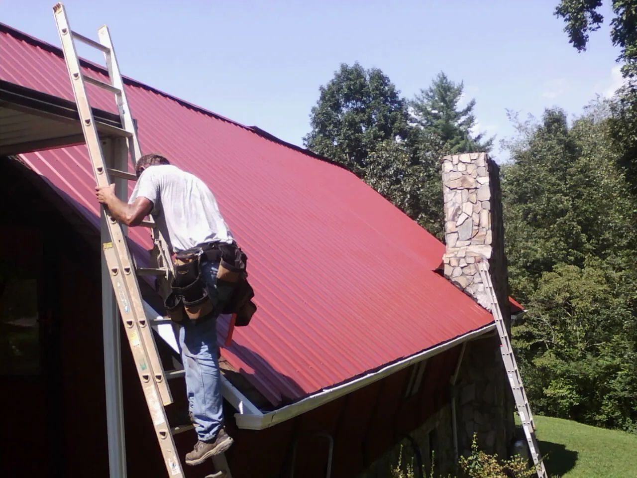 Roofer on a ladder beside a red metal roof and stone chimney — Soffit & Fascia Repair by Roofing Brothers in Houlka